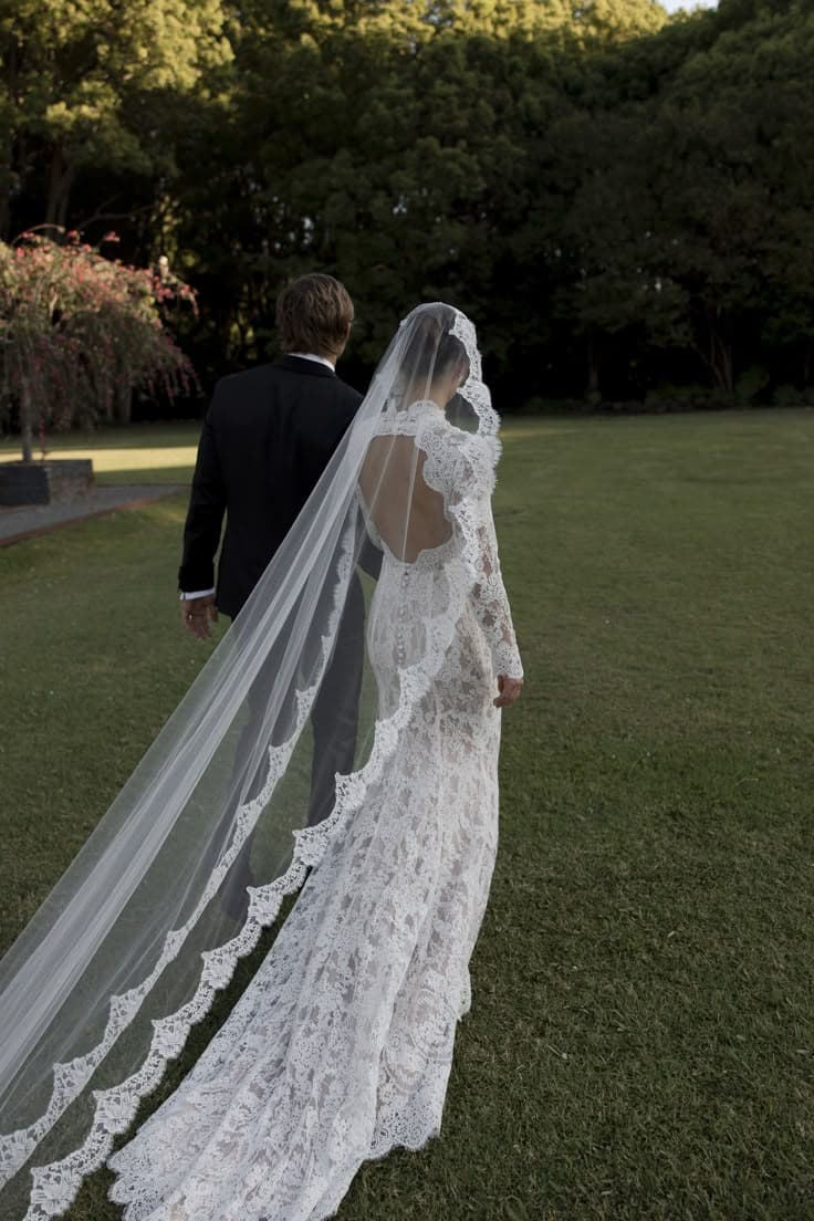 A bride from behind in a lace gown and long cathedral veil on a lawn