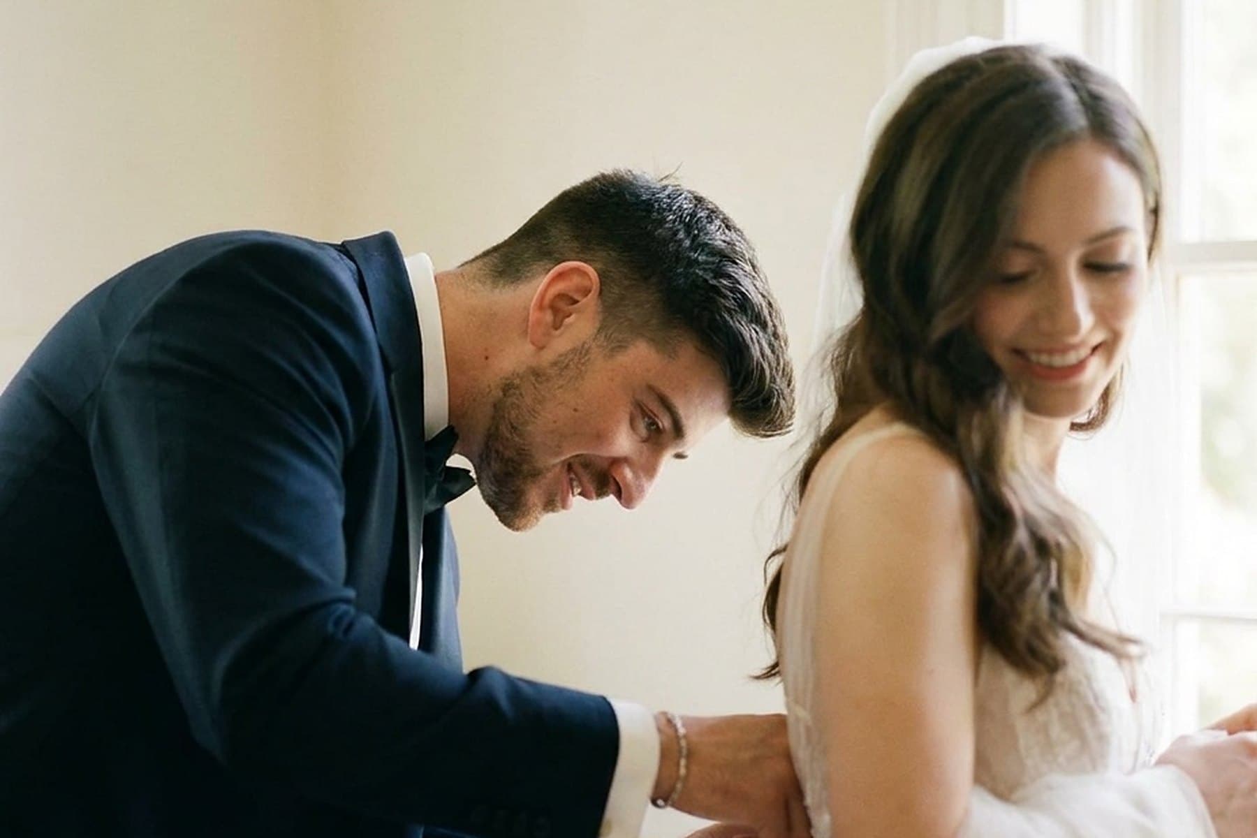 A groom fastening a bride's dress in a sunlit room