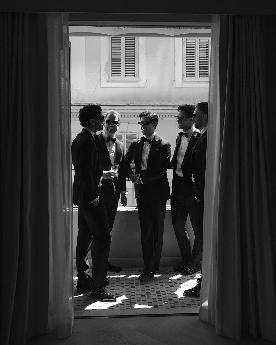 Groomsmen in black tuxedos laughing by a window