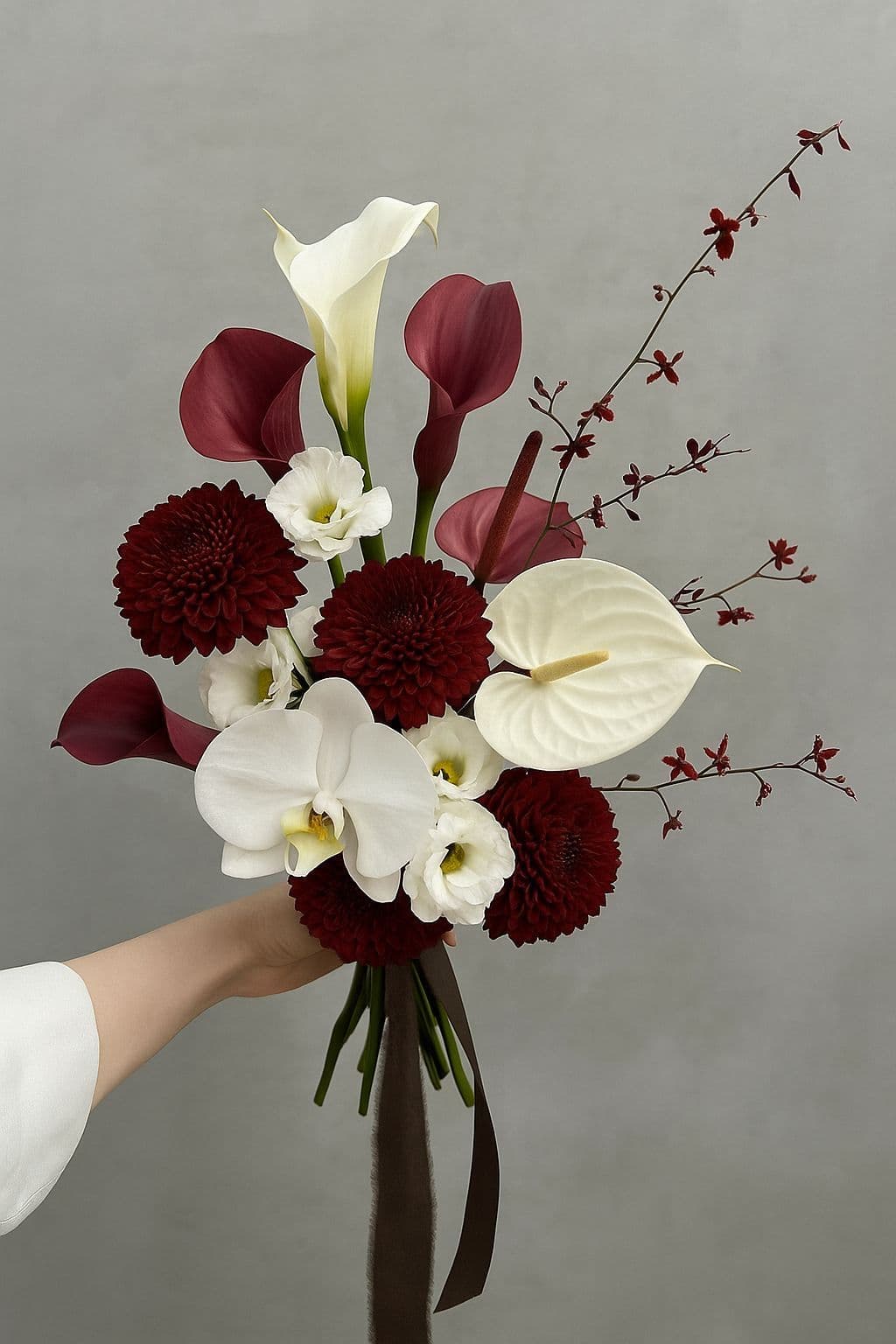 A bouquet of burgundy dahlias, calla lilies and white orchids held against a plain grey wall