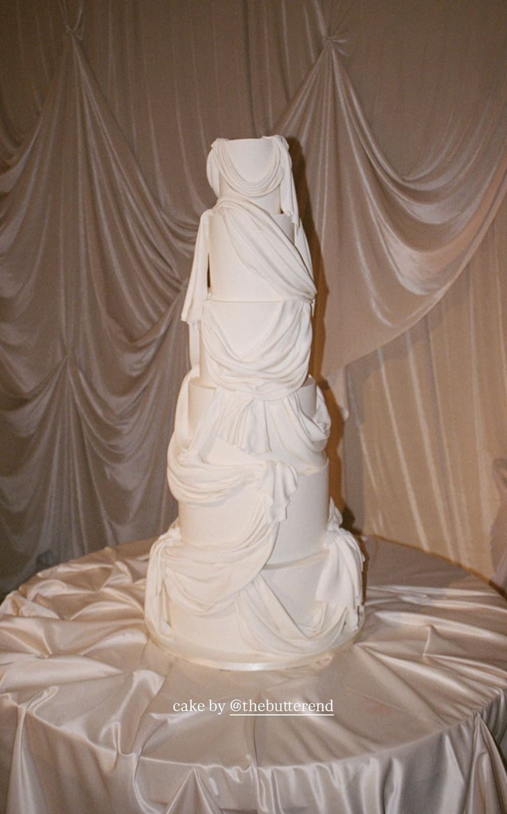 A tall five-tier white wedding cake with fondant draped and gathered across every tier, in front of draped curtains