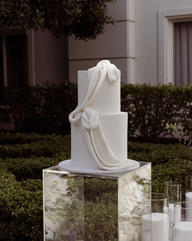 A two-tier white wedding cake wrapped in a single sweep of draped fondant, on a mirrored plinth