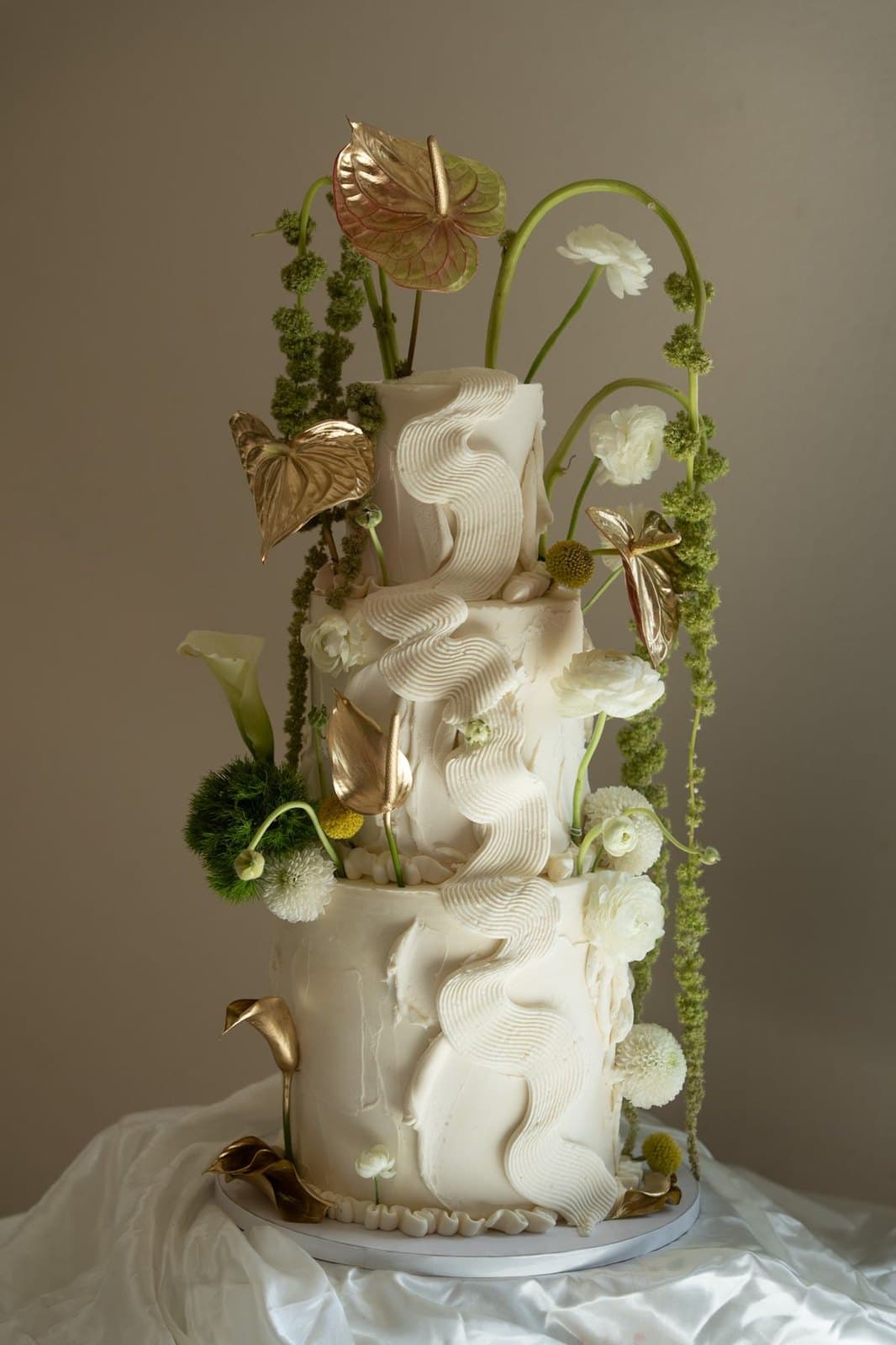 A three-tier textured white wedding cake with a piped ruffle running down the front, gilded anthuriums, ranunculus and trailing green amaranthus