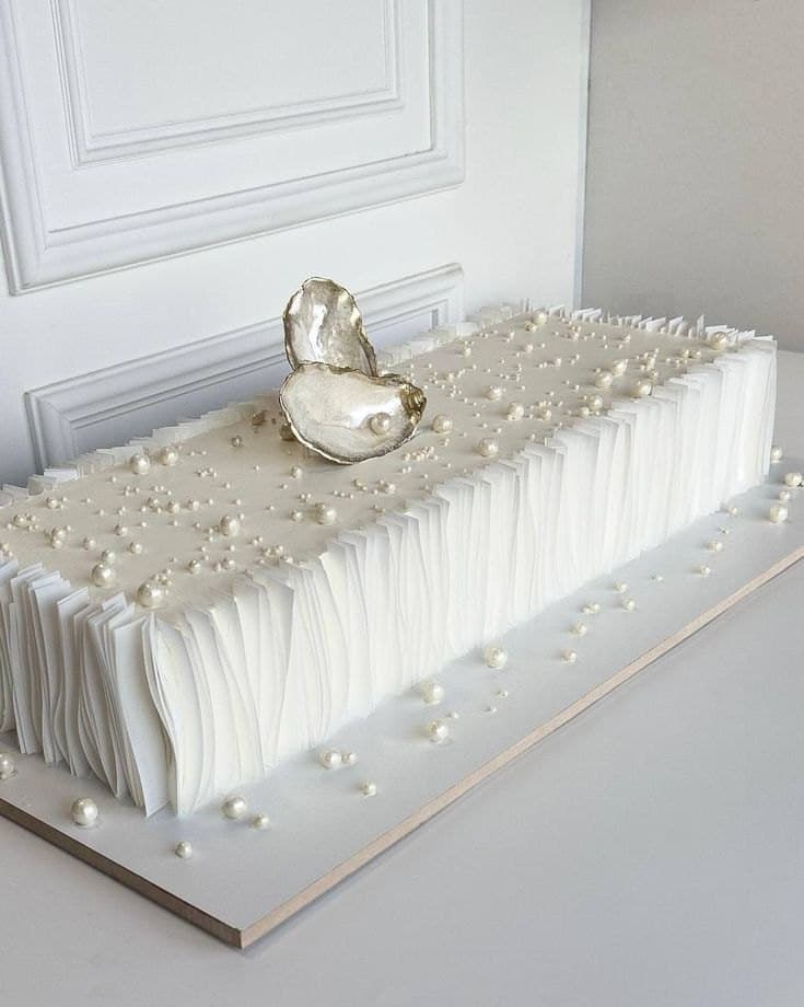 A rectangular white wedding cake with pleated wafer-paper sides, scattered sugar pearls and a single gilded oyster shell on top