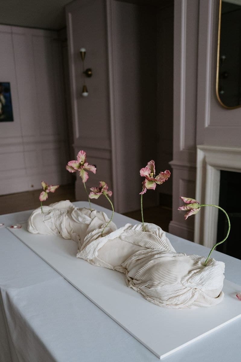 A low, sculptural wedding cake made of pleated and folded cream fondant, with pink sugar tulips rising from it, on a white table in a mauve room