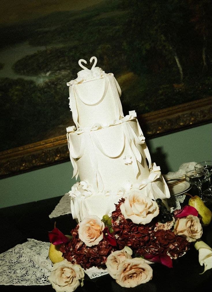 A three-tier white wedding cake with piped ribbon swags and bows and two swans on top, on a lace cloth with roses and hydrangea at the base, in front of an oil painting