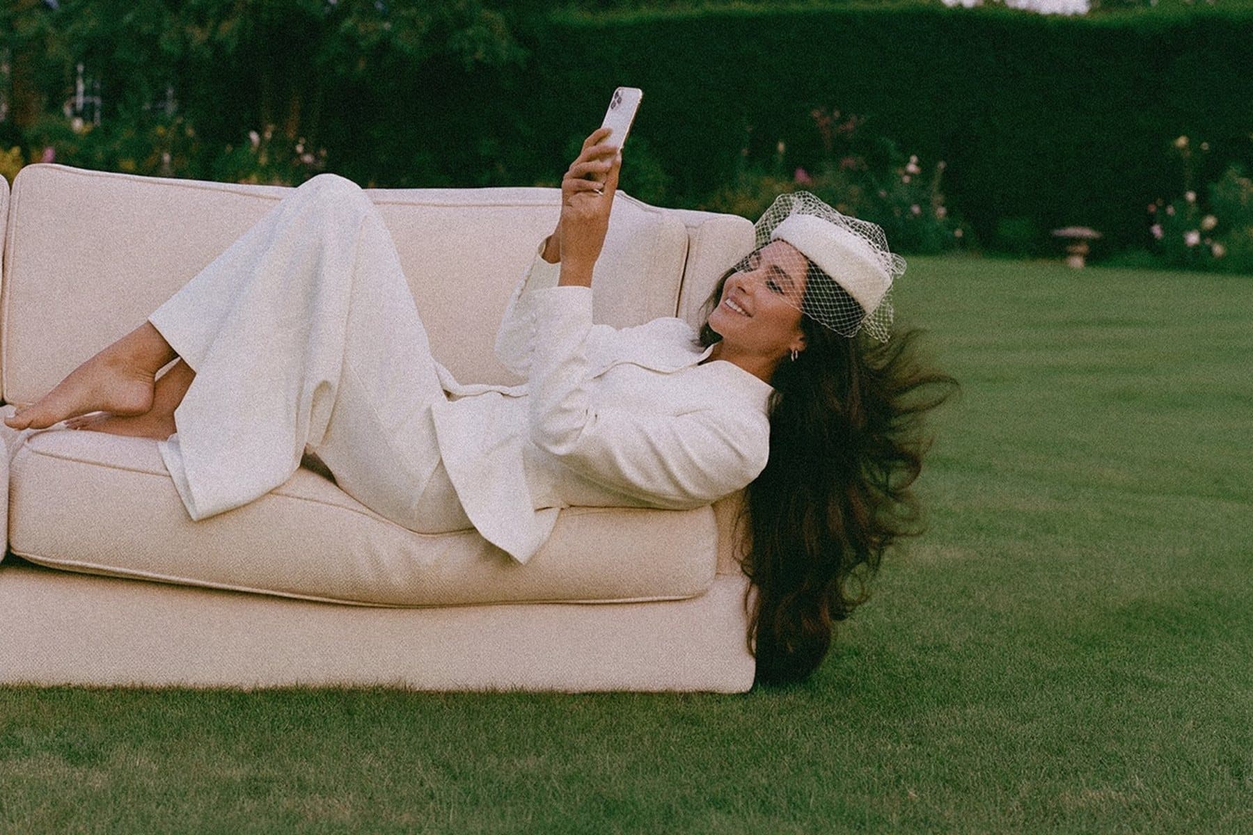 A bride in a white suit and veiled hat lying on a sofa on a lawn, reading her phone