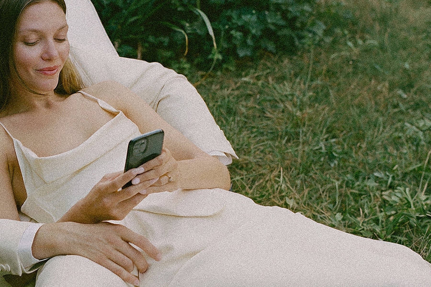 A bride in a cream slip dress reading her phone, resting against her partner on the grass