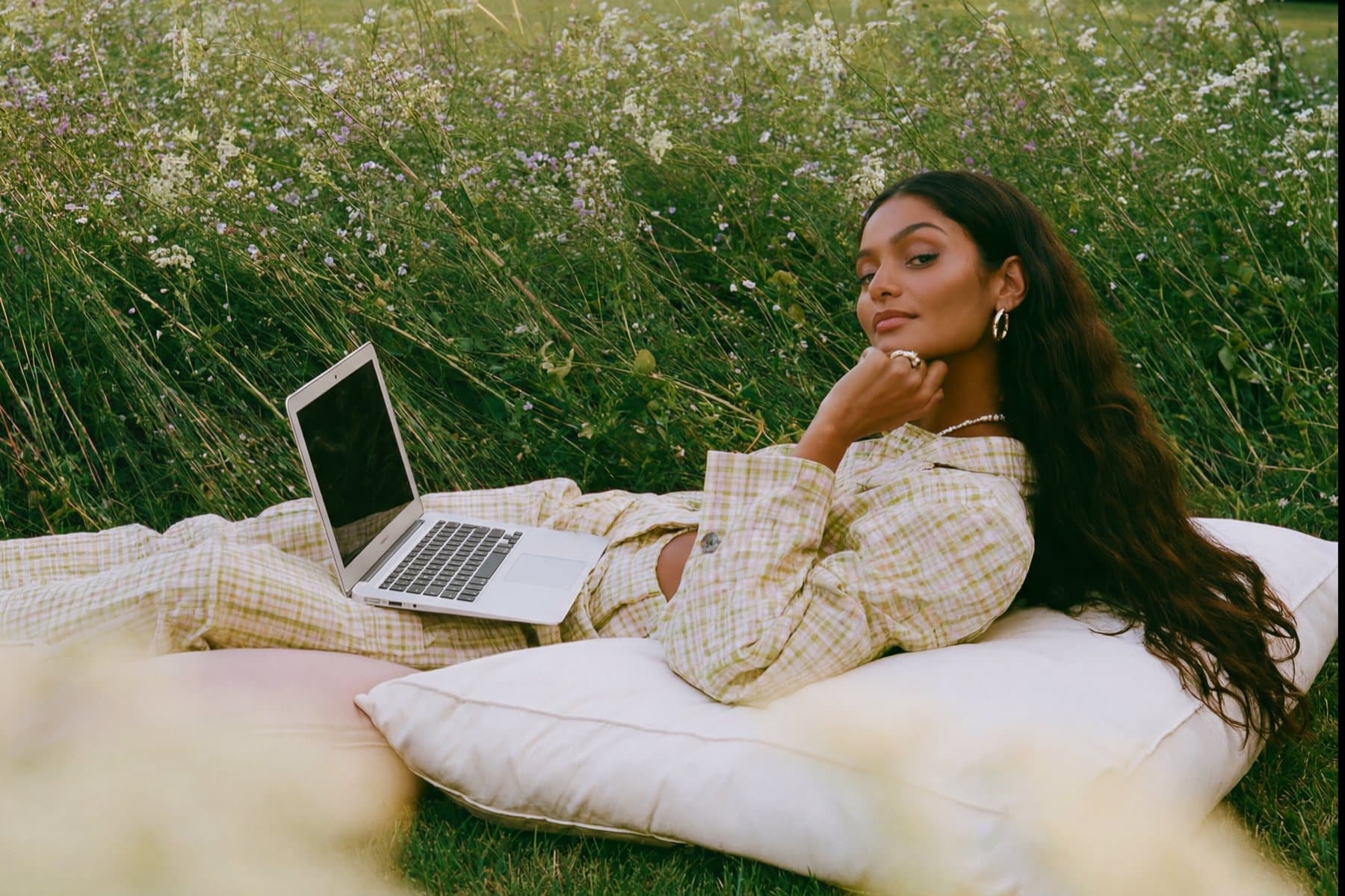 A woman in a checked suit lying on cushions in a wildflower meadow beside an open laptop