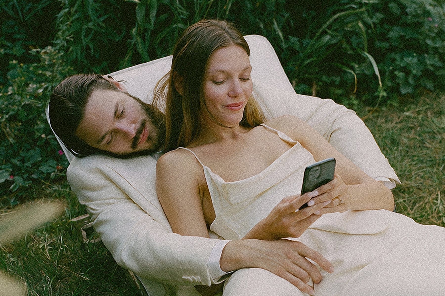 A couple in cream lying in the grass, the bride reading her phone