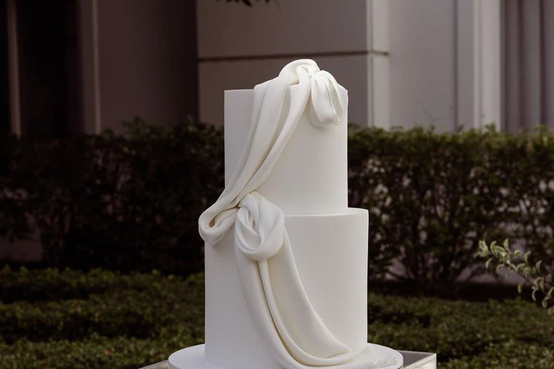 A two-tier white wedding cake with draped fondant on a mirrored plinth in a garden