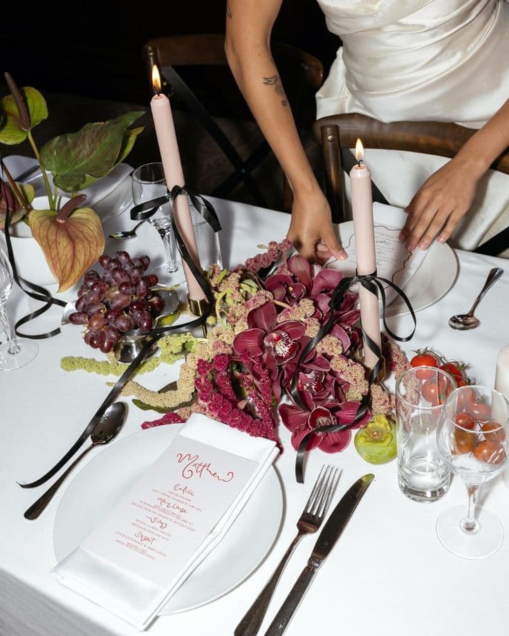 A place setting with a handwritten menu card, tapered candles, grapes and burgundy orchids laid low along the table